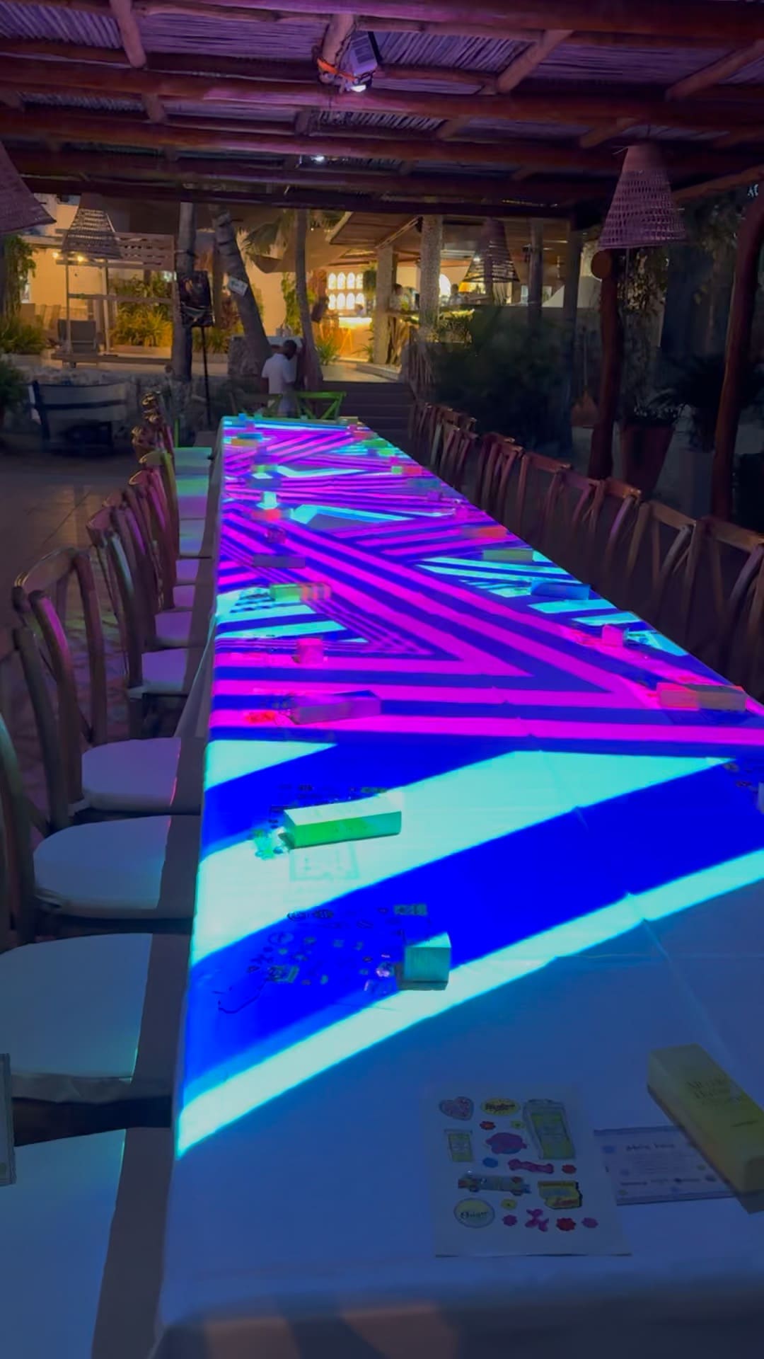 Mesa larga en la playa con proyección de geometría neón magenta y azul, sillas de madera y techo de palma